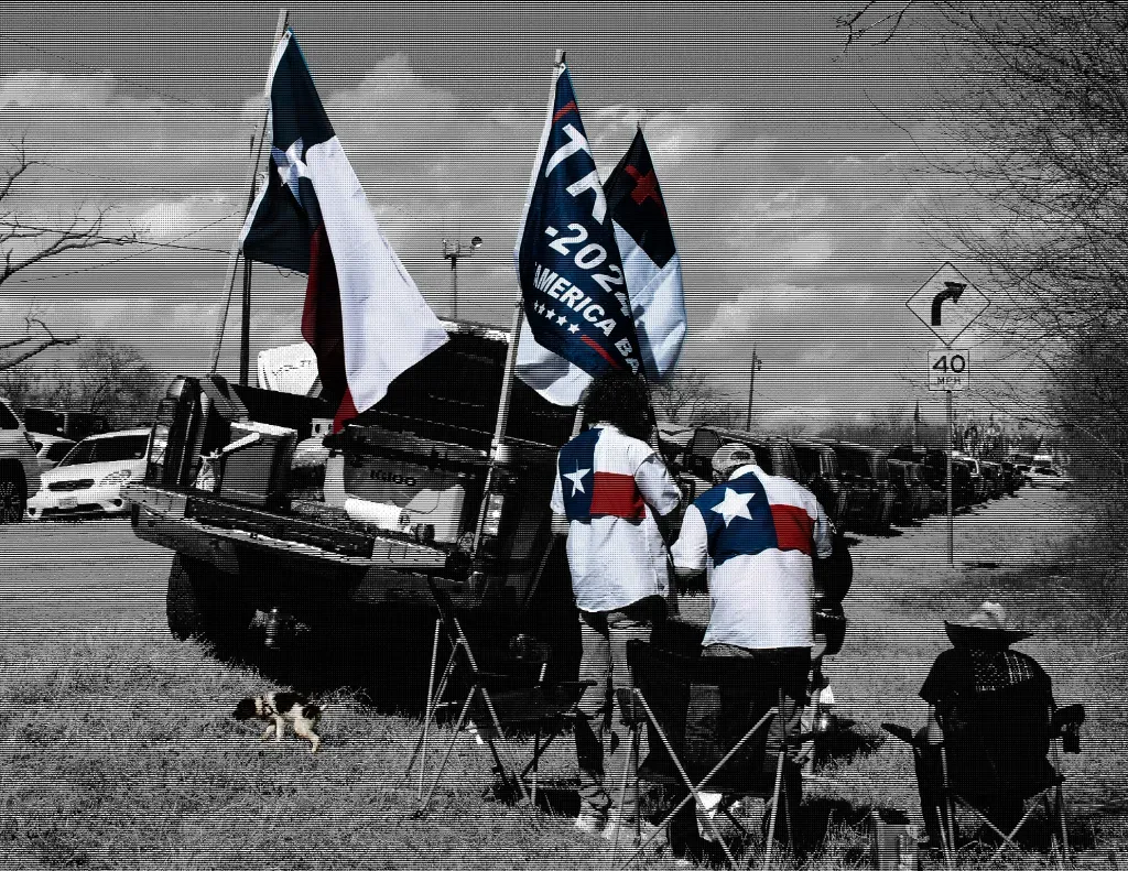 a black and white photo (with reds and blues visible) of people at the rally having a BBQ near their truck which has multiple flags including a Trump and Christian nationalist one..